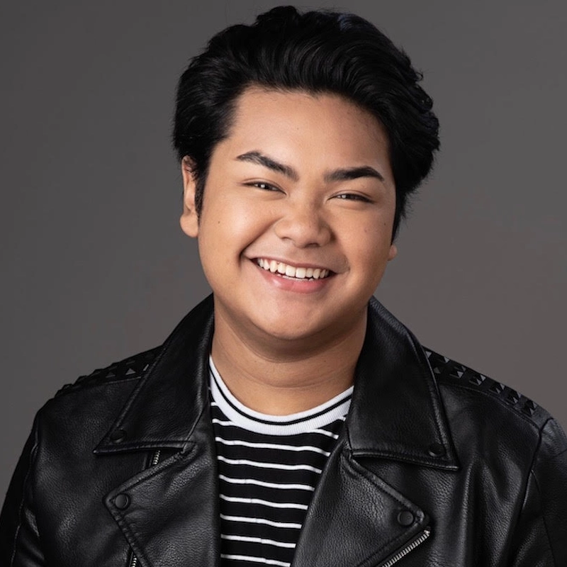 A young person with short black hair smiles at the camera. They wear a black leather jacket over a black-and-white striped shirt. The background is gray and plain, highlighting their warm, friendly expression.