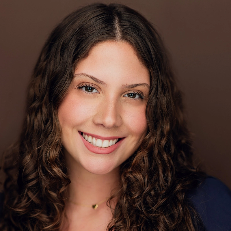 Young woman with long, curly brown hair smiles at the camera. She has light skin, blue eyes, and is wearing a dark top. The background is plain brown, and the lighting is soft, highlighting Ireyssa Cardona’s natural features.