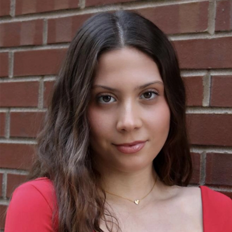Sara-Louise Oakes, a young woman with long brown hair and fair skin, stands in front of a red brick wall. She wears a red top with a scoop neckline and a delicate gold necklace with a small heart pendant, looking at the camera with a slight smile.