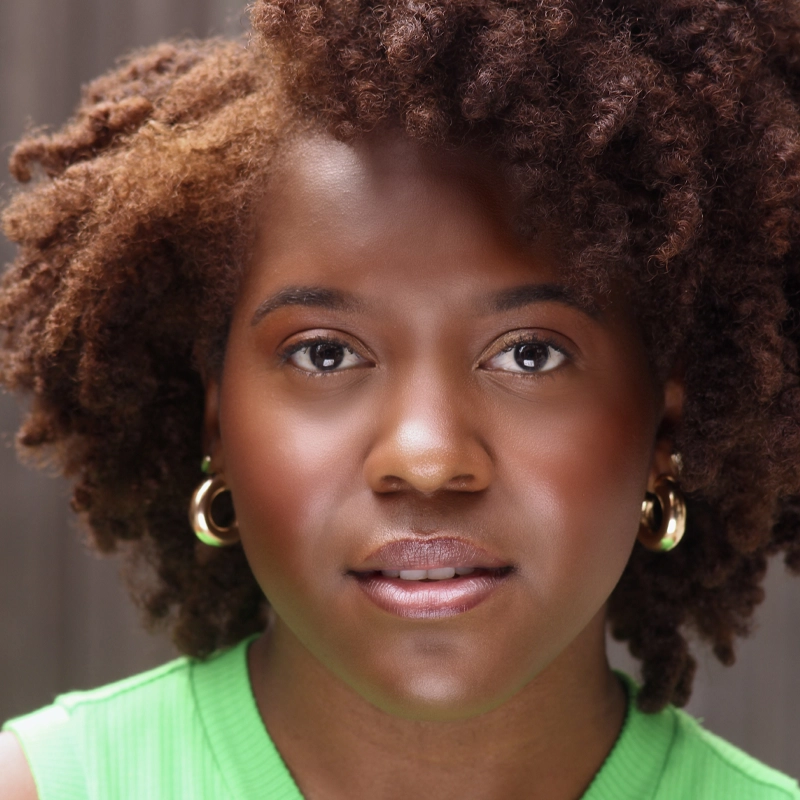 A woman with natural curly hair, identified as Jalyn Crosby, wears gold hoop earrings and a bright green top. She faces the camera with a calm expression, the softly blurred background highlighting her glowing complexion and features.