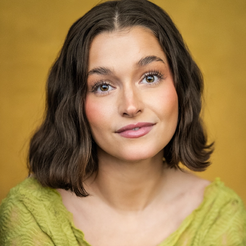 A young woman with short wavy brown hair and hazel eyes looks at the camera, smiling slightly. She wears natural makeup and a textured light green top. The background is a warm mustard yellow, creating a soft and inviting portrait.
