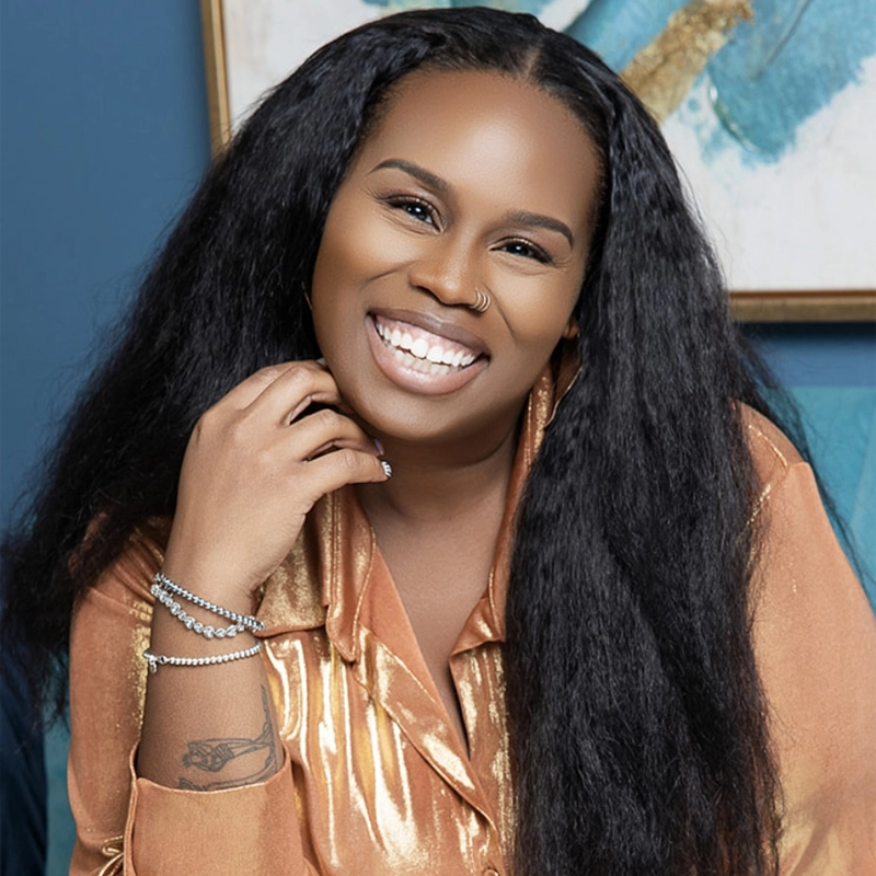 Desirée Dabney, a smiling woman with long, straight black hair, wears a shiny gold satin blouse. She rests her hand near her chin, displaying bracelets and a tattoo on her wrist. The background features a blue wall and abstract art.