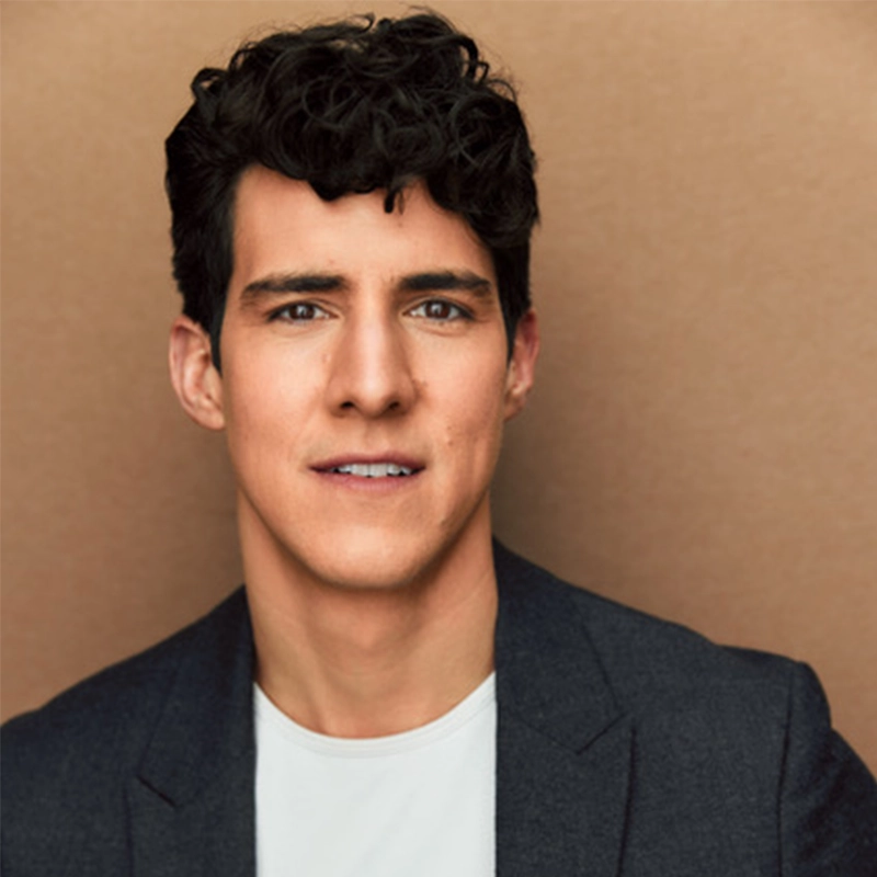 Daniel Arana, a young man with wavy dark hair, fair skin, and brown eyes, is wearing a dark blazer over a white shirt. He faces the camera with a slight smile against a plain, light brown background.