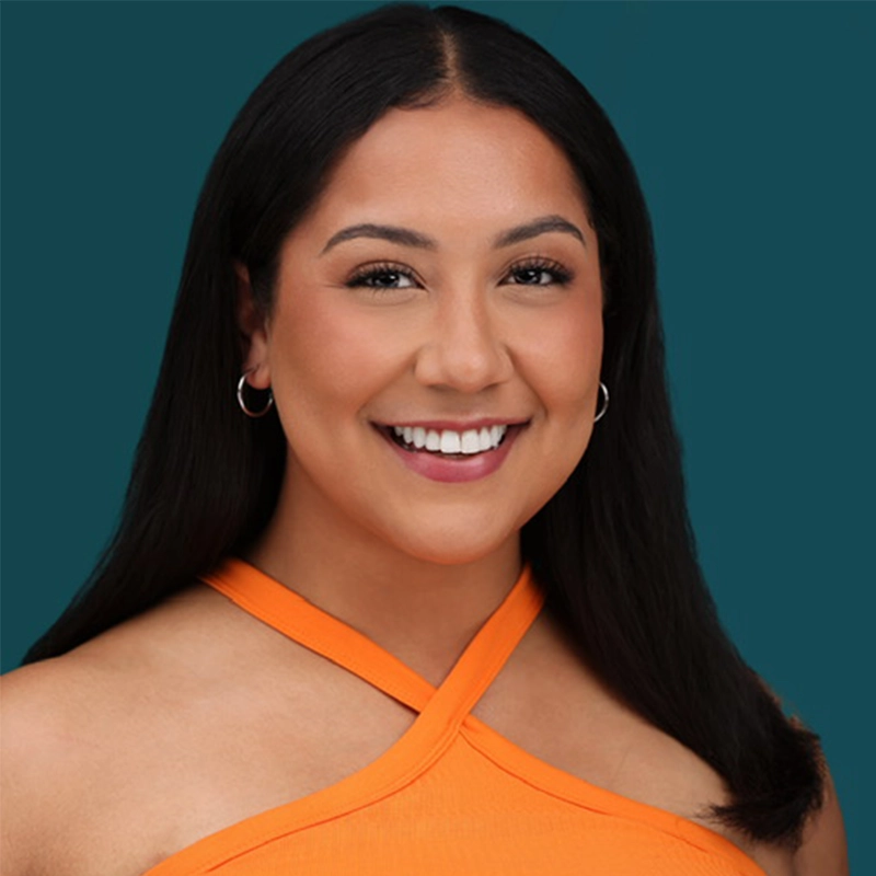 Melissa Aliotta smiles at the camera, her long, straight dark hair framing her face. She wears hoop earrings and a bright orange, sleeveless top with crisscross straps against a solid teal background.