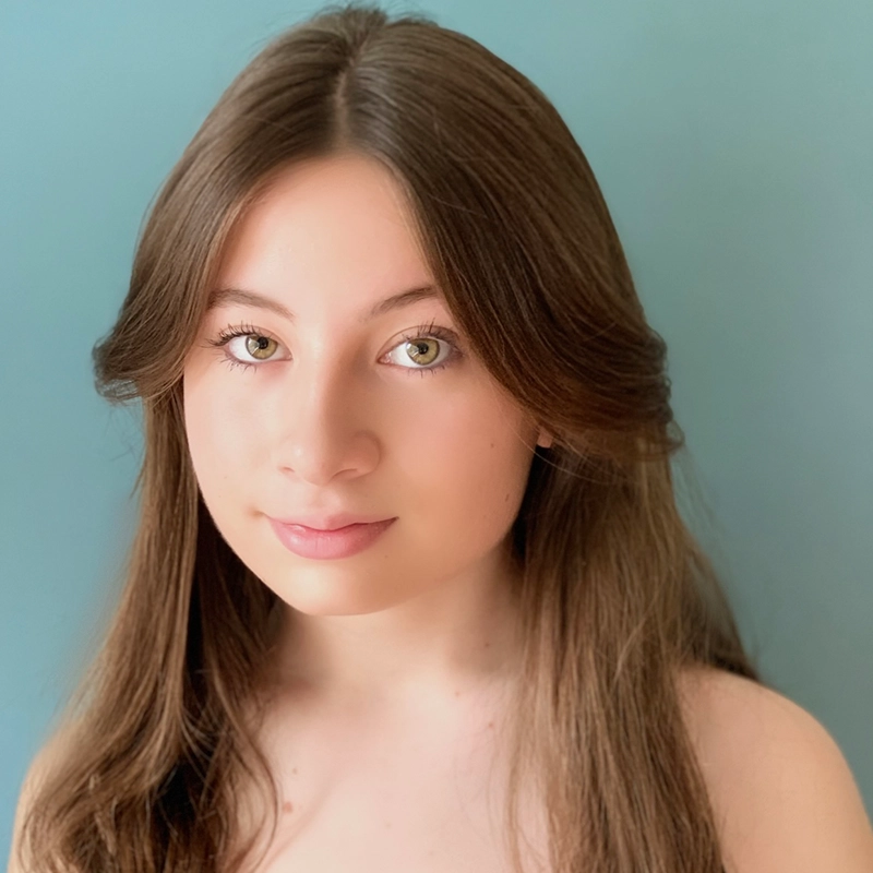 A young woman with long, straight brown hair and fair skin faces the camera against a plain light blue background. Reminiscent of a Killian Allgeier portrait, she has hazel eyes, subtle makeup, and a calm expression; her center-parted hair softly frames her face.