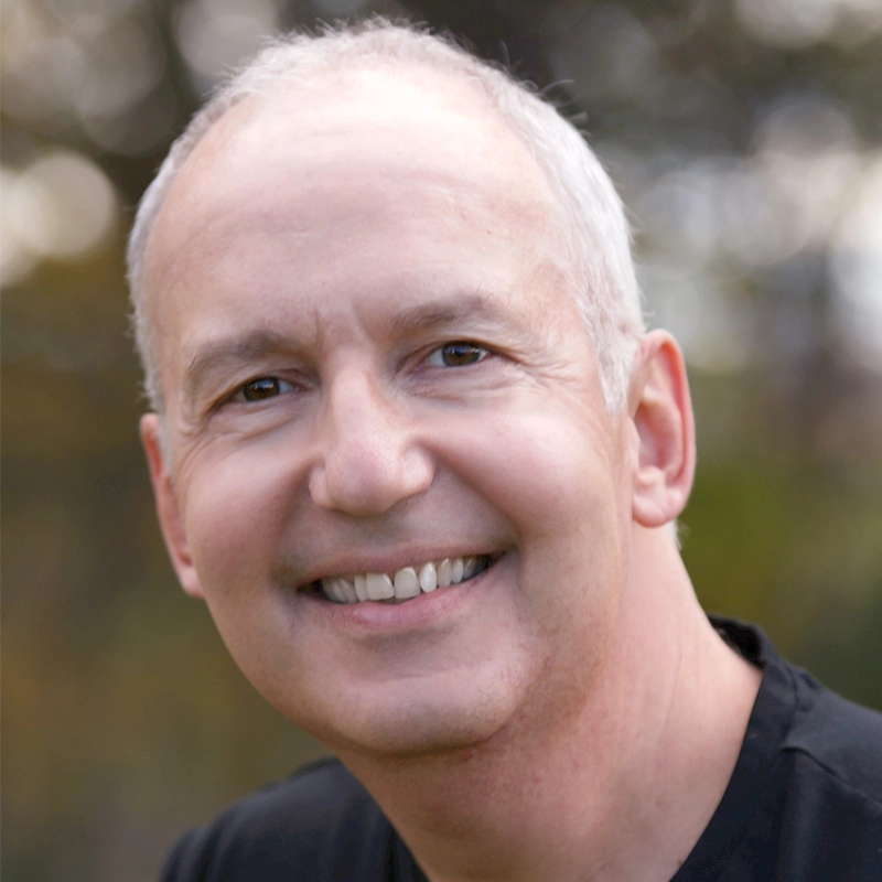A middle-aged man with short gray hair, David Lowenstein, smiles warmly at the camera. He is wearing a black shirt and is outdoors, with soft-focus trees and greenery in the background. Natural lighting highlights his cheerful expression.