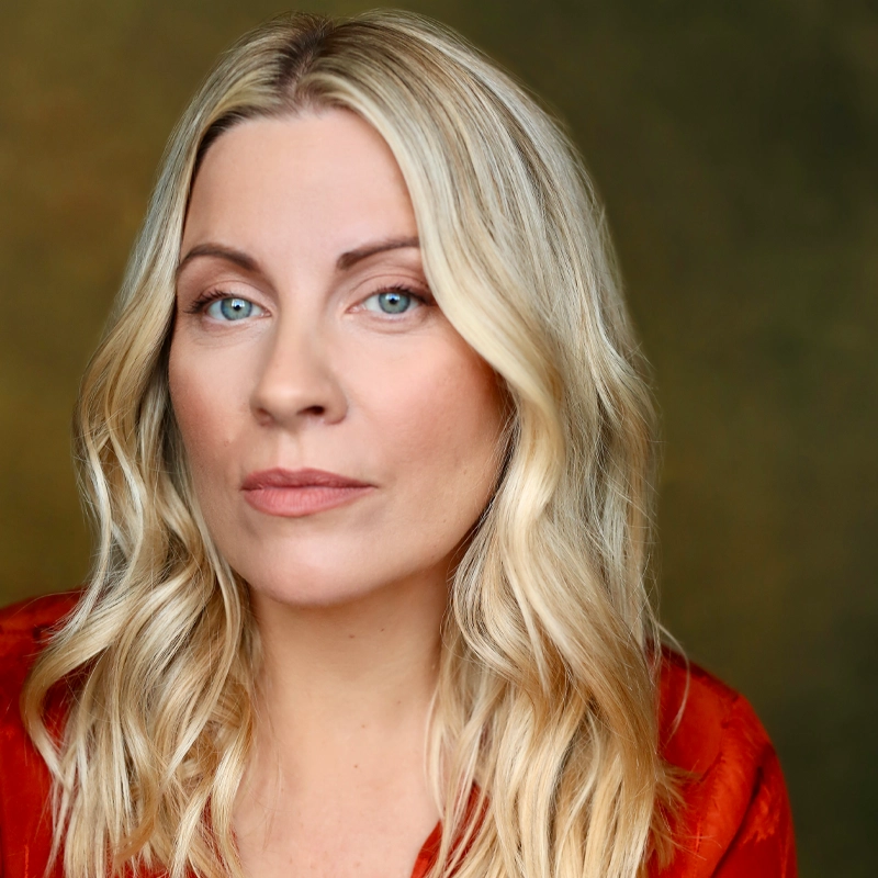 A woman with long, wavy blonde hair and blue eyes looks at the camera with a neutral expression. Reminiscent of West End musical theatre star Louise Dearman, she wears a red top; the background is a soft, muted green and brown gradient.