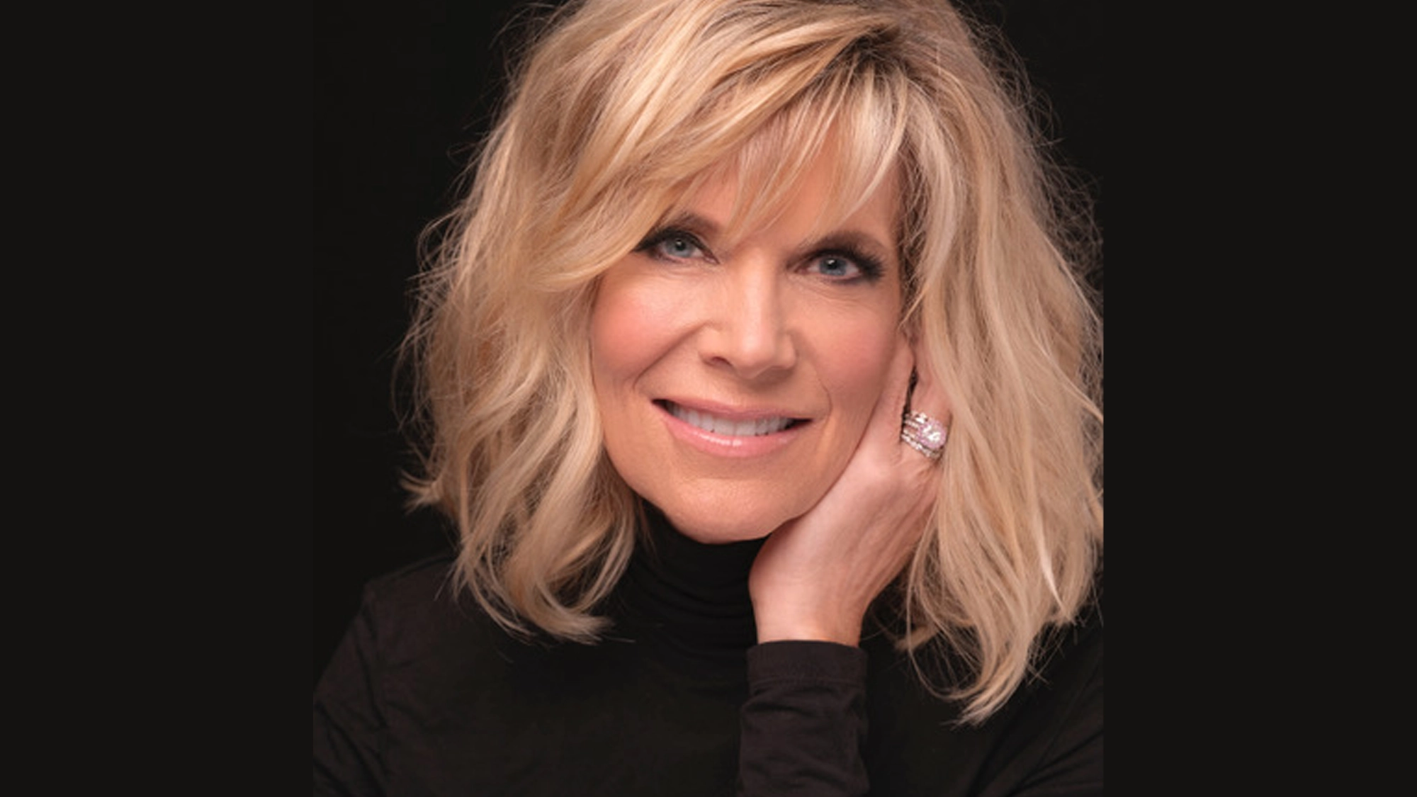 A smiling woman with shoulder-length blonde hair and side-swept bangs rests her chin on her hand. Reminiscent of Debby Boone, she wears a black turtleneck and a large ring, the dark background highlighting her face and hair—it's been a while since such charm shone through.