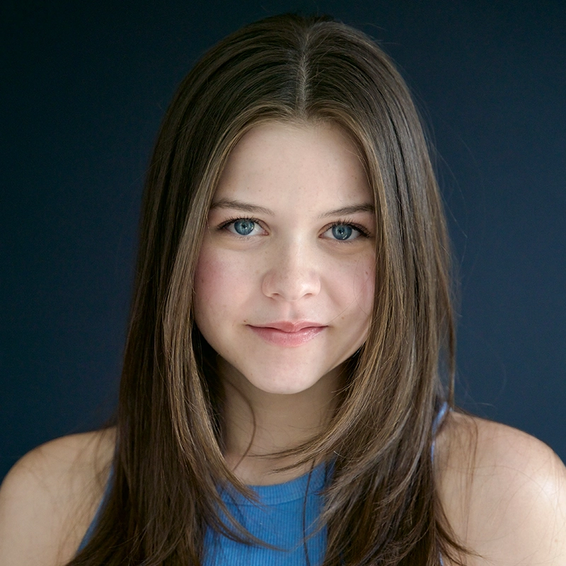 A young person with long straight brown hair and light blue eyes smiles softly at the camera. Wearing a sleeveless blue top, they are posed against a dark Kodiak blue background. Their complexion is fair, and their expression remains calm and friendly.