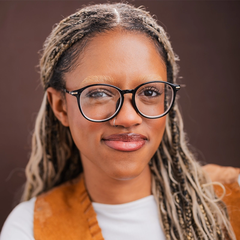 A young person with light brown skin, long braided hair with blonde highlights, large black glasses, and a nose ring smiles slightly. They wear a white shirt under a brown vest. The background is a plain dark brown. This portrait captures Ireyssa Cardona’s distinctive style and quiet confidence.