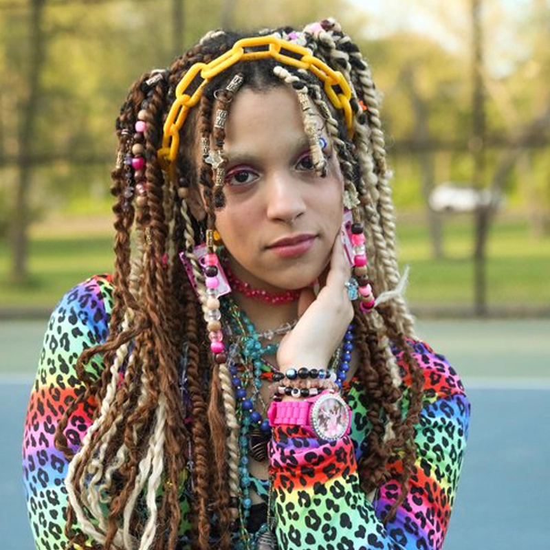 Chiane Ekweonu poses outdoors with long, beaded braids adorned with colorful beads and a yellow hair accessory. Sporting a vibrant leopard-print top, pink watch, and layered necklaces, she stands against a blurred park and tennis court backdrop.