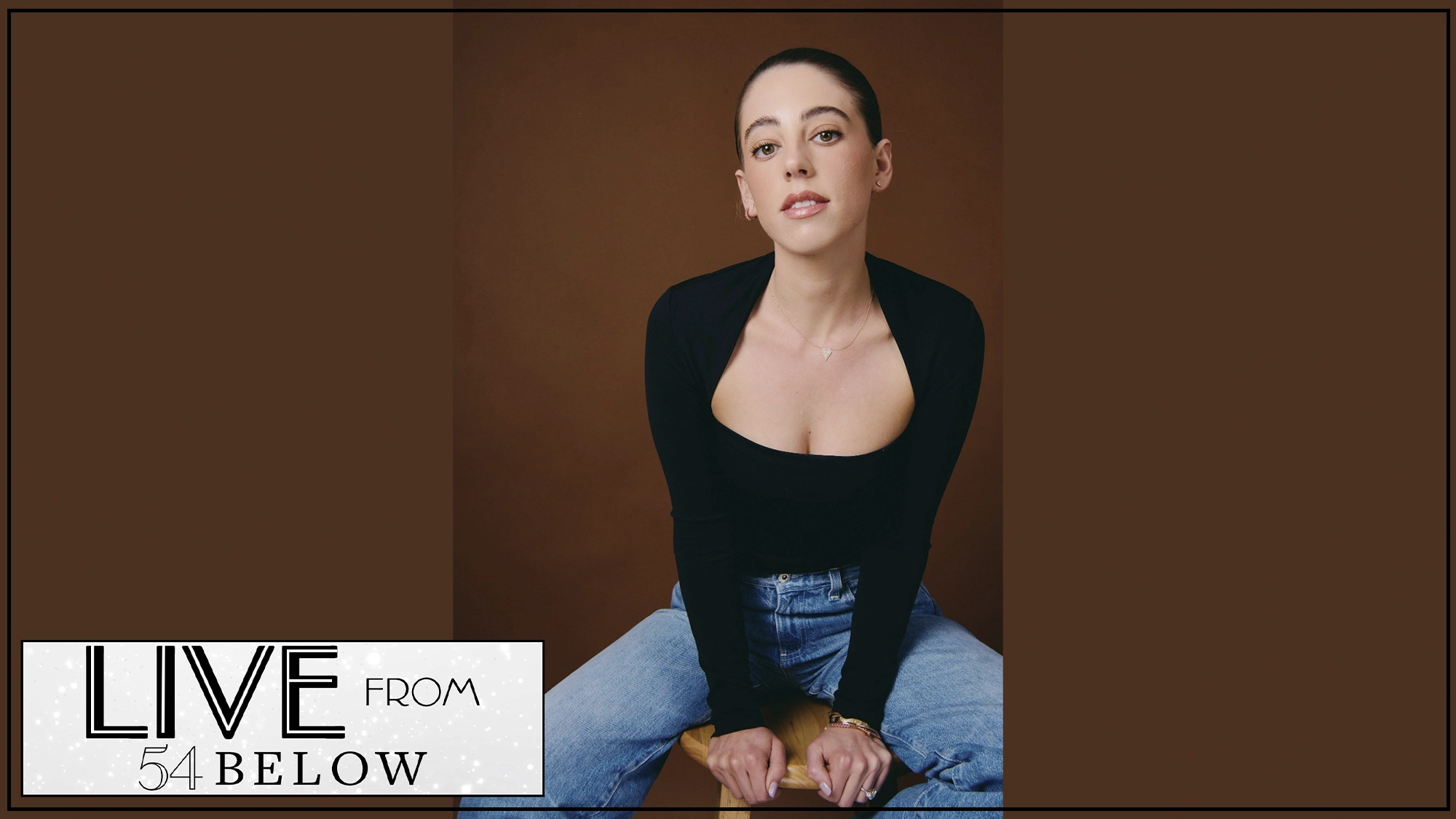 A woman with dark hair pulled back sits on a stool against a brown background. She wears a black scoop-neck top and blue jeans. In the lower-left corner, a 