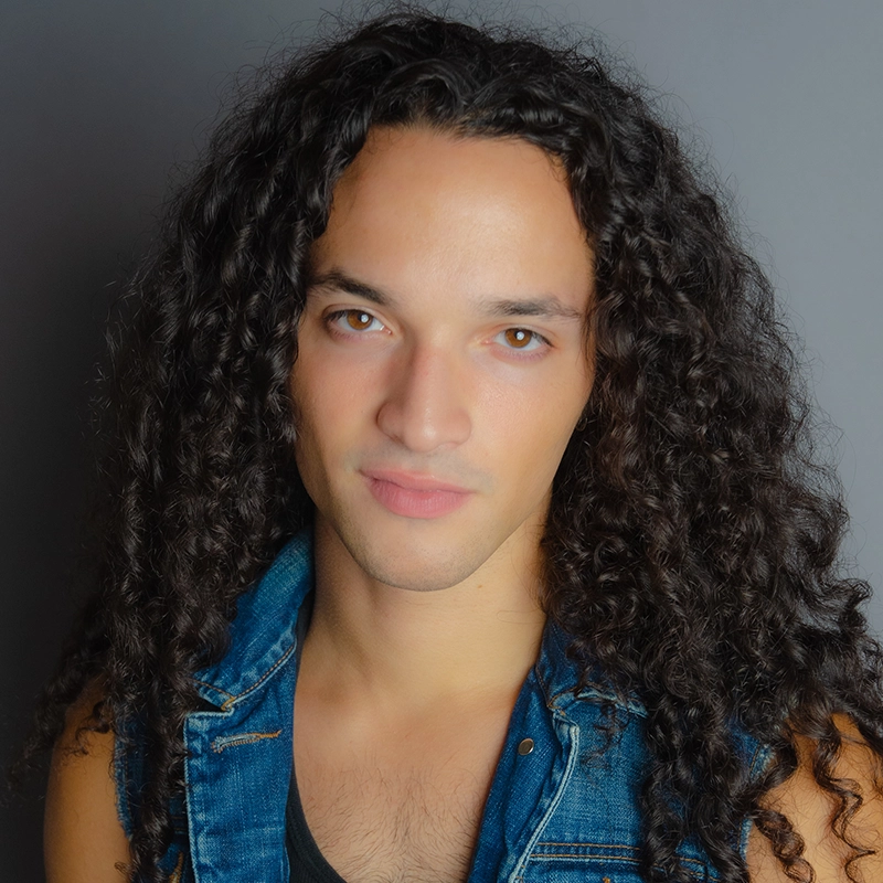 Katie Yeomans, with long, dark, curly hair and light skin, faces the camera against a gray background. She wears a sleeveless denim vest over a black shirt and has a calm, neutral expression.