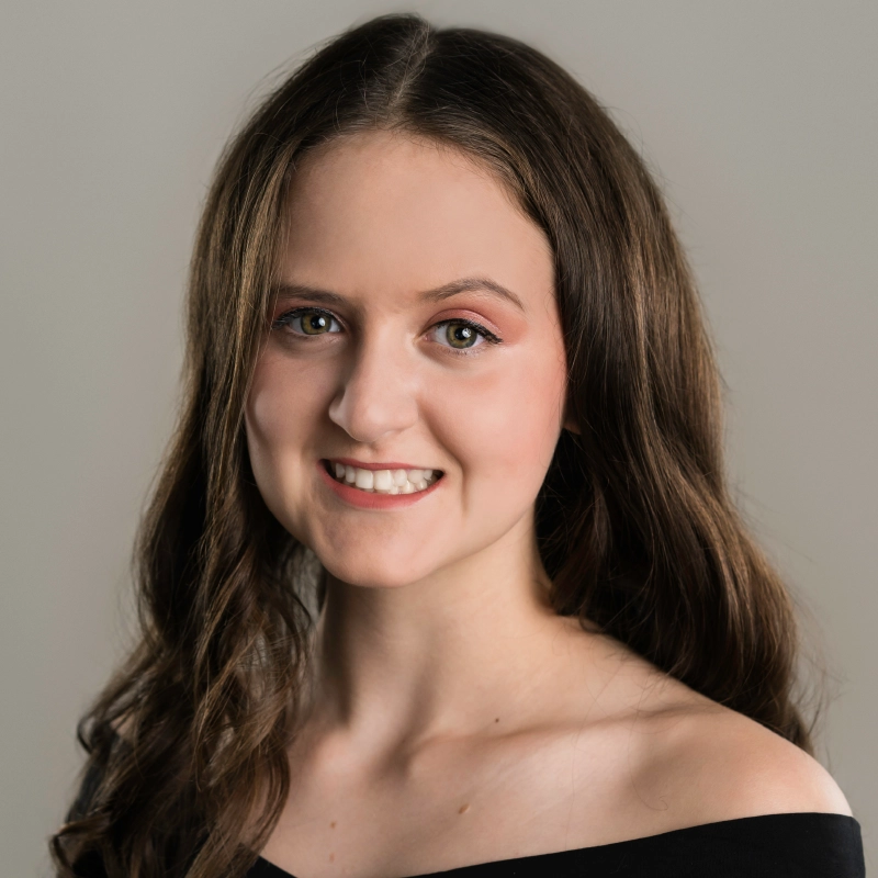 Lyla Karekinian, a young woman with long, wavy brown hair and green eyes, smiles at the camera. She wears natural makeup and a black off-the-shoulder top against a plain light gray background for a simple, professional portrait.