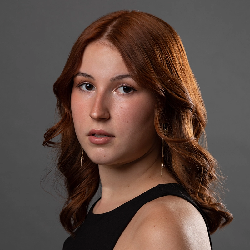 Lyla Karekinian, a young woman with wavy, reddish-brown hair, poses against a neutral gray background. She wears a sleeveless black top and long, dangling earrings, her expression neutral as she looks slightly past the camera with soft makeup.