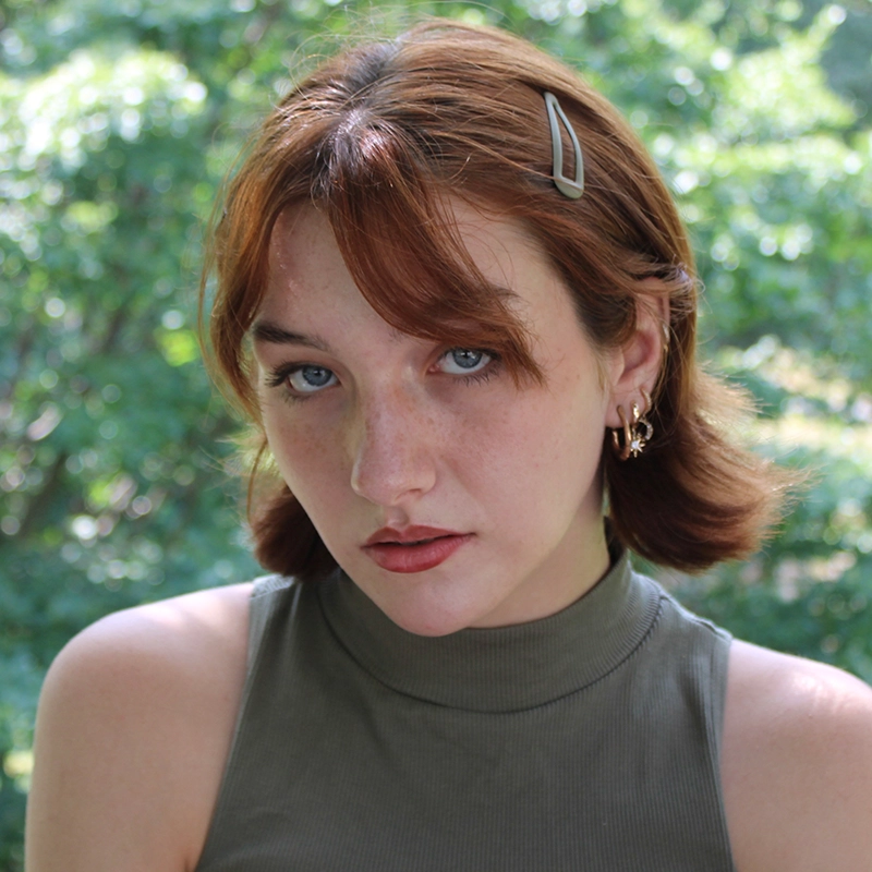 A young woman with short auburn hair, wearing a silver hair clip and gold earrings, looks at the camera with a neutral expression. Dressed in a sleeveless, high-neck olive green top, she stands amid green foliage in soft focus—a portrait by Kodiak Thompson.