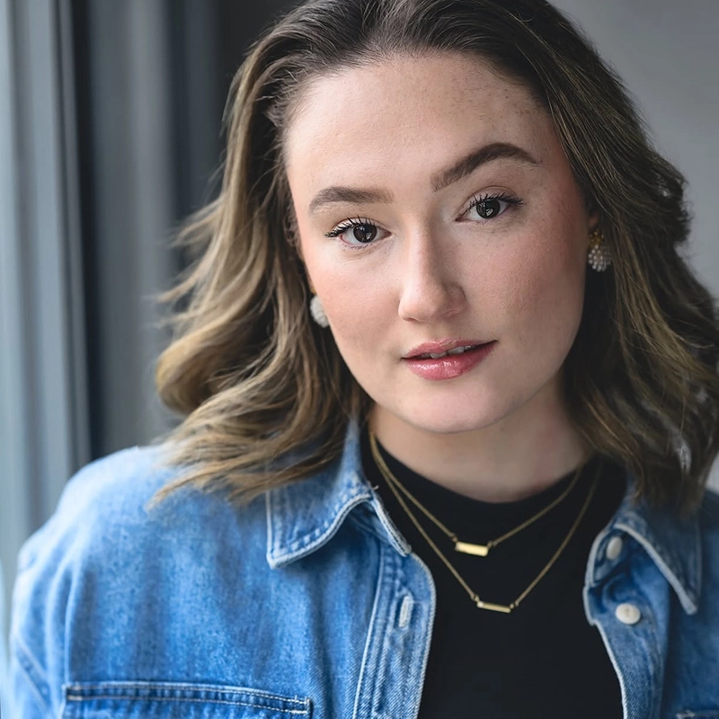 A young woman with wavy brown hair wears a denim jacket over a black top and layered gold necklaces. She has natural makeup, stud earrings, and looks confidently at the camera while standing next to a window with soft daylight.