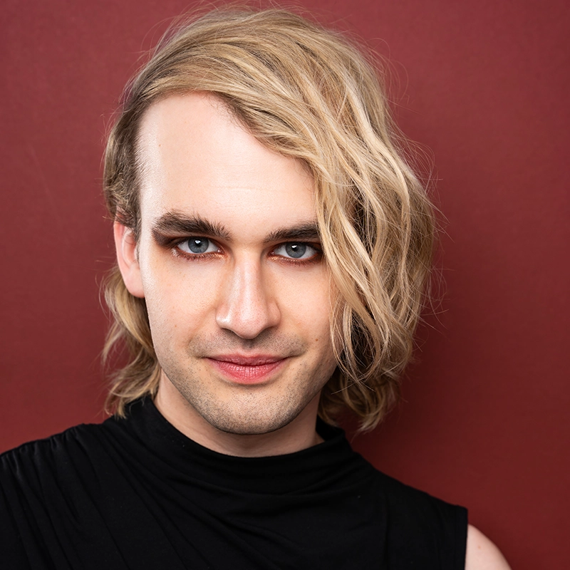 A person with fair skin and shoulder-length wavy blonde hair is looking confidently at the camera. They have bold eye makeup and wear a sleeveless black top. The background is a solid deep red color.