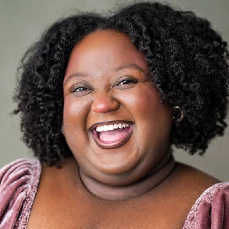 A smiling person with dark curly hair wearing a mauve velvet top. Their face is joyful, mouth open in laughter, showing teeth. The background is neutral and softly blurred, keeping full focus on the person’s expressive, happy face.