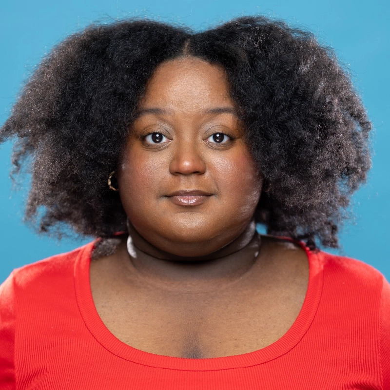 A person with medium-dark skin and natural, voluminous black hair looks directly at the camera. They wear a red scoop-neck top and a black choker, set against a plain blue background. Their expression is neutral and confident.