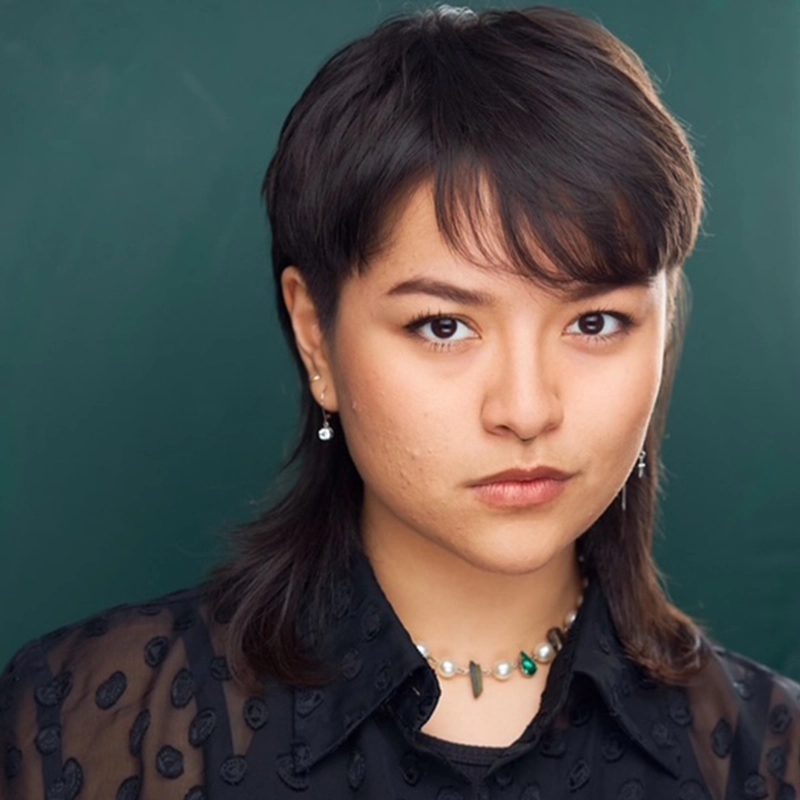 A person with straight, dark hair in a mullet style—reminiscent of Kourtnie Dwornikowski—wears a sheer black shirt with textured dots, a beaded necklace, and drop earrings, looking directly at the camera against a solid dark green background.