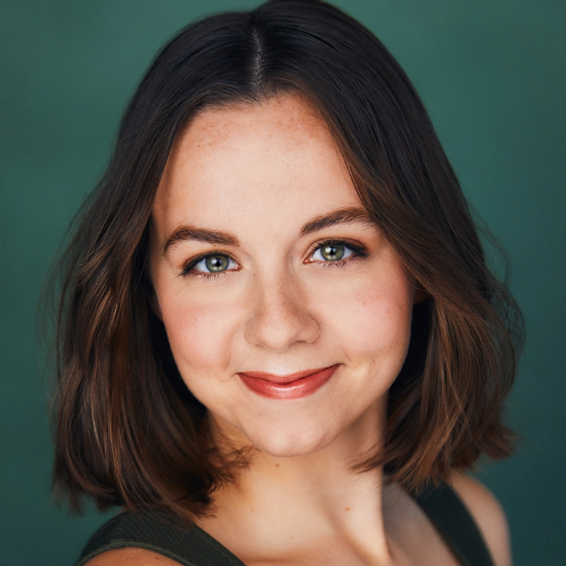 Katelynn Fahrer, a young woman with fair skin and brown, shoulder-length hair, smiles softly. She has green eyes, neatly groomed brows, natural makeup with pink lipstick, and wears a sleeveless dark top against a muted green background.