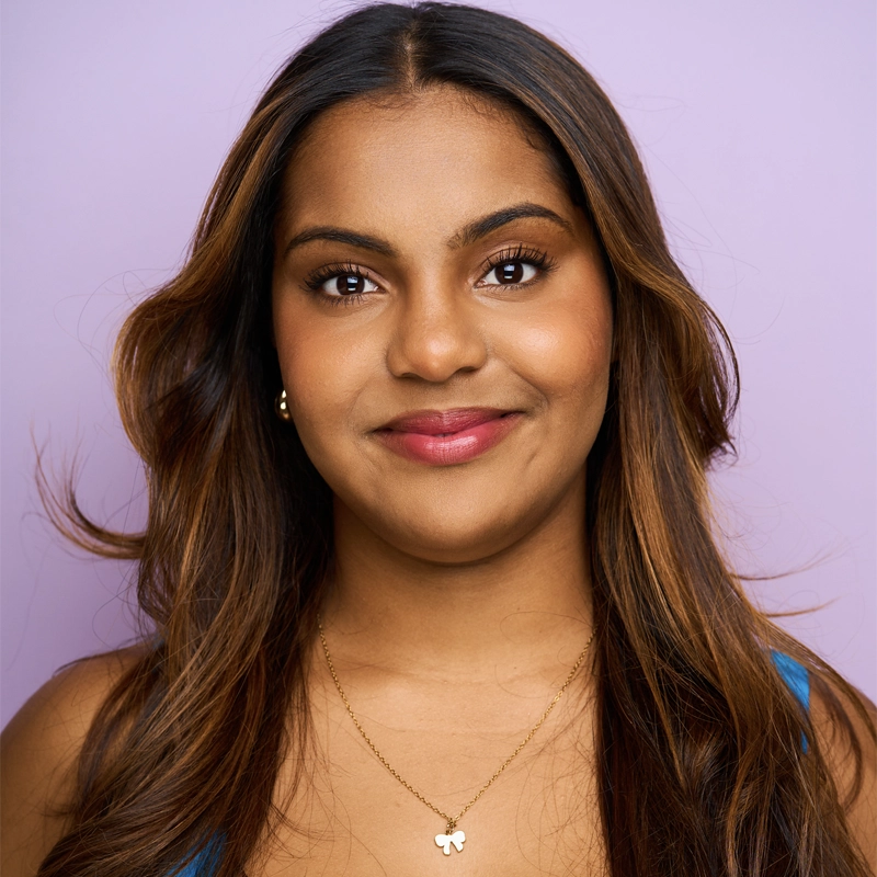 Naiya Fernandez, a woman with medium brown skin and long, wavy brown hair, smiles gently at the camera. She wears a blue top, gold stud earrings, and a gold butterfly pendant necklace against a solid light purple background.