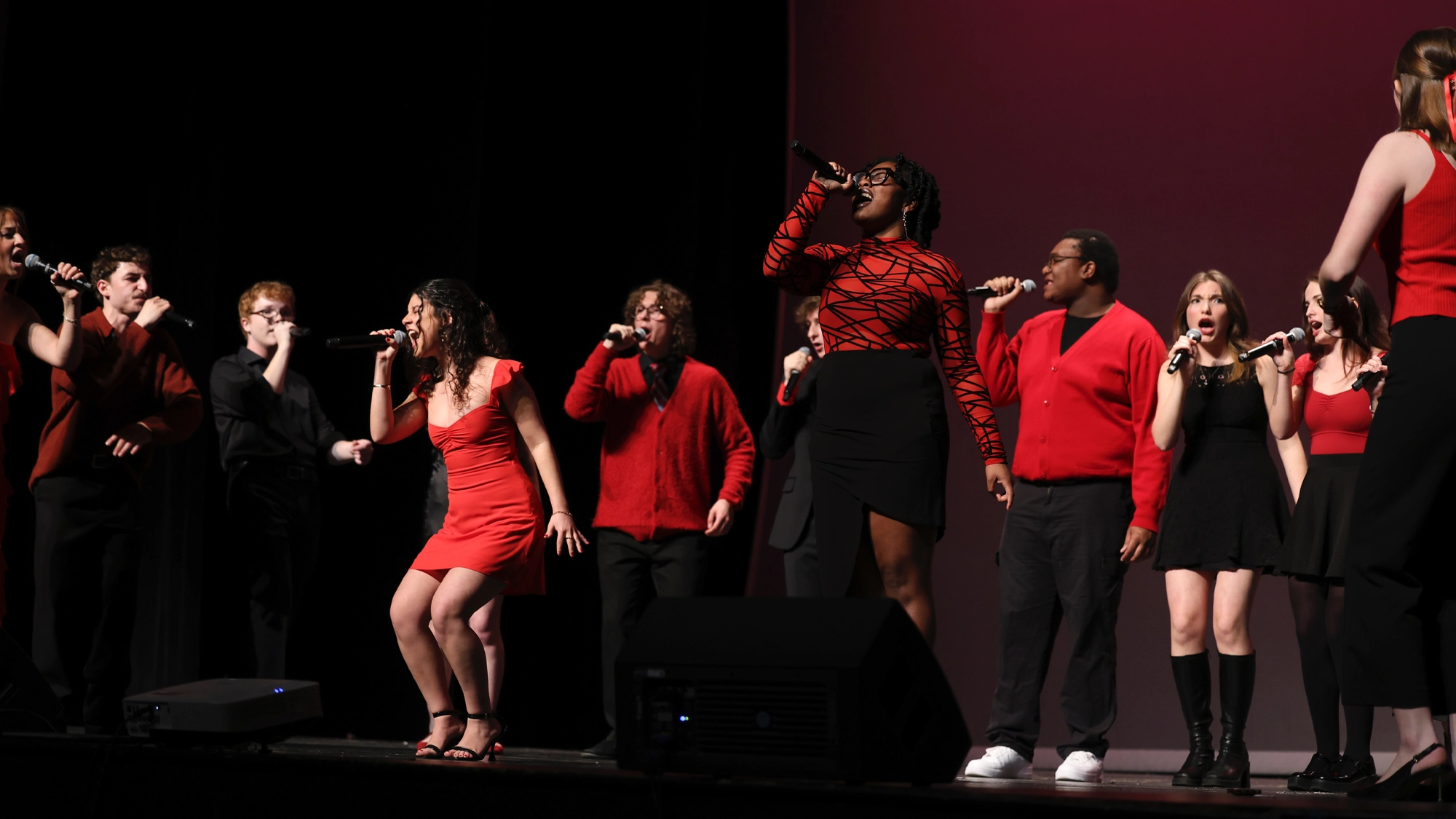 Nine singers from Fordham University’s Hot Notes perform on stage, all holding microphones. They wear coordinated red and black outfits, with a dark background and a pinkish-purple spotlight highlighting two energetic vocalists in the foreground.