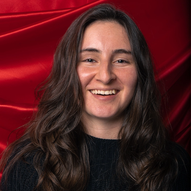 Francesca Pastore, a young woman with long, wavy brown hair and fair skin, smiles at the camera. She wears a black sweater and stands before a rich red satin backdrop, giving the portrait a warm and inviting feel.