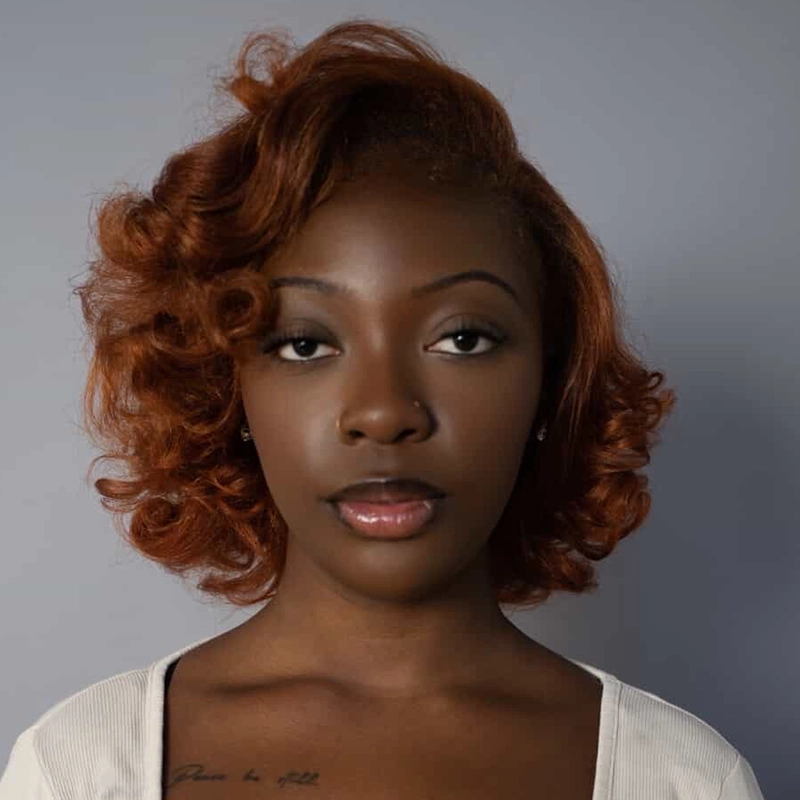 Lyla Karekinian, a young Black woman with smooth, medium-dark skin and auburn, wavy hair, gazes calmly at the camera. She wears neutral makeup, a white top, and small earrings against a plain, soft gray background, exuding quiet confidence.