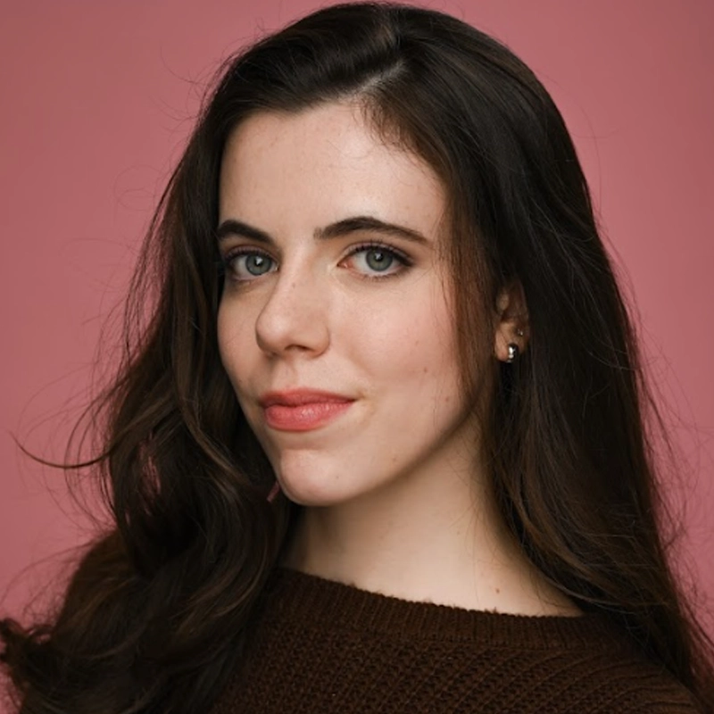 Renata Gabella, a young woman with long, wavy brown hair and light eyes, poses against a pink background. She wears a brown knit top and small stud earrings, looking at the camera with a slight, confident smile.