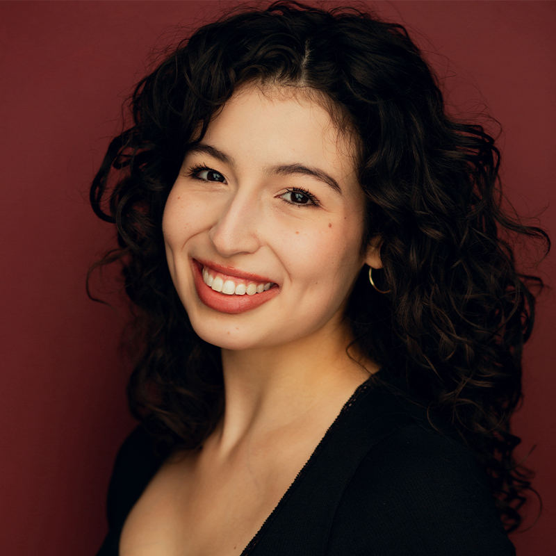 Gabriela Barrett, a young woman with curly dark hair, smiles warmly at the camera. She wears a black top, small hoop earrings, and natural makeup. The solid maroon background gives her portrait a professional and friendly appearance.