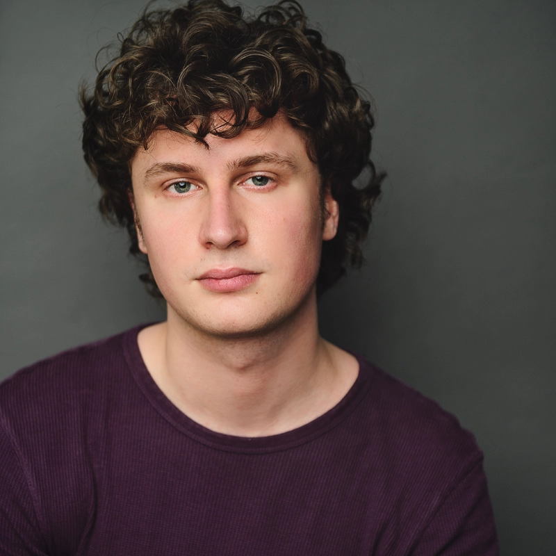 Ethan Garrepy, a young man with curly brown hair and fair skin, wears a purple ribbed shirt. He looks directly at the camera with a neutral expression against a plain dark gray background, making him the clear focus of the image.