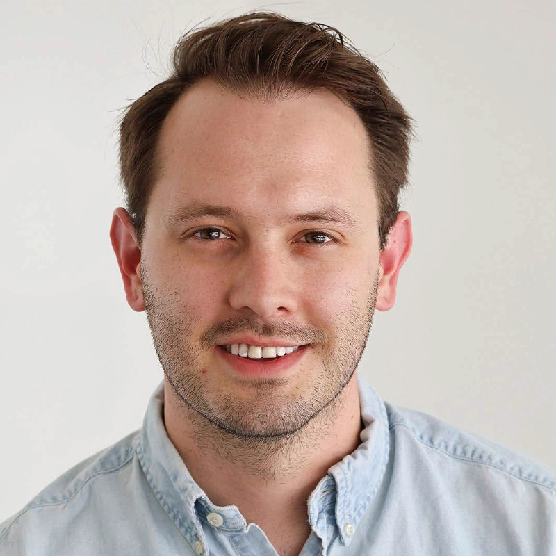 A man with short brown hair and light stubble smiles at the camera. He is wearing a light blue button-up shirt, posed against a plain background, radiating a friendly, approachable vibe—perfect for a professional profile like Katie Krasovec’s.