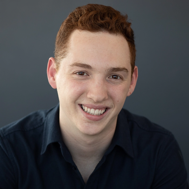 A young man with short, curly red hair, Nicholas Kraft, smiles warmly at the camera. He wears a dark collared shirt against a plain dark gray background. His head is slightly tilted, and he appears relaxed and approachable.
