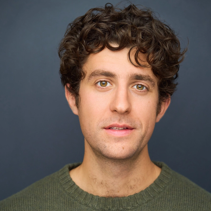 Caleb Graham, a young man with curly brown hair and hazel eyes, wears an olive green sweater. He looks directly at the camera with a neutral expression against a plain, dark blue-gray background under soft, even lighting.