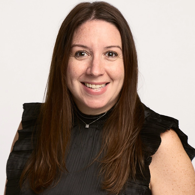 Pam Schmeir Hacker, a woman with long brown hair and fair skin, smiles at the camera. She wears a sleeveless black top with ruffled shoulders and a small necklace. The plain white background gives her portrait a professional and friendly appearance.