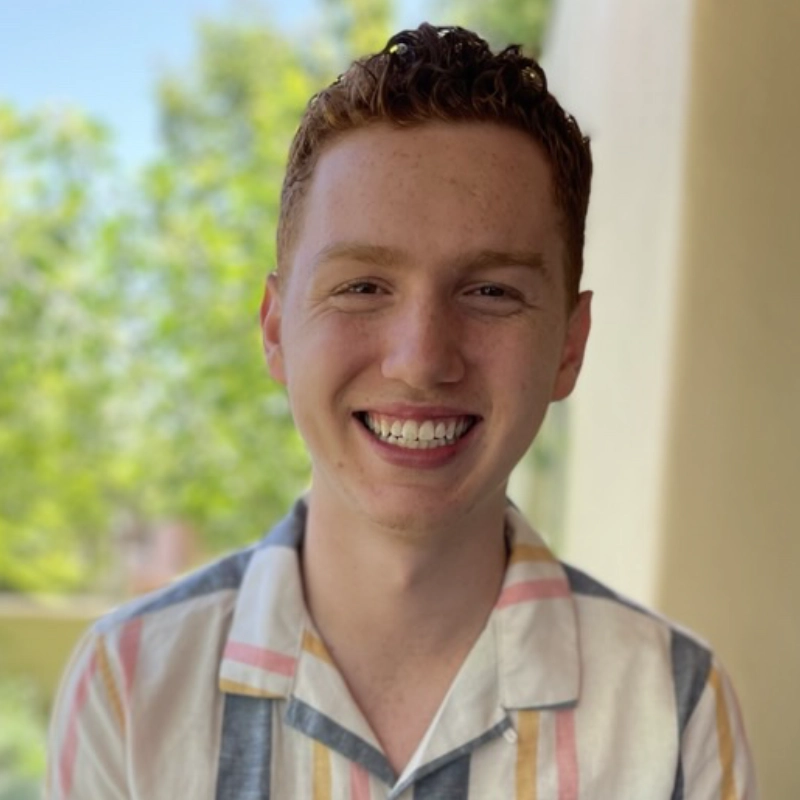 Nicholas Kraft, a young man with short curly red hair, smiles widely, showing his teeth. He wears a light-colored, short-sleeve shirt with pastel stripes. The softly blurred green trees and sunlit wall suggest he is outdoors on a bright day.