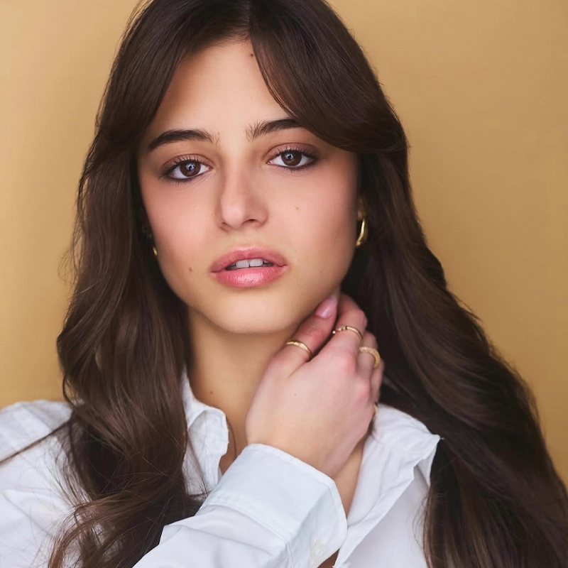 A young woman with long brown hair, wearing a white shirt and gold hoop earrings, poses against a solid tan background. She gazes forward with a neutral expression, her hand gently touching her neck, and her makeup is natural and minimal—embodying the understated elegance often seen in Eve Rosenstein’s signature style.