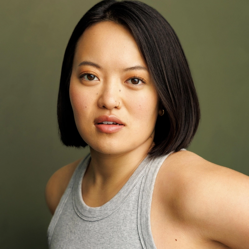 A person with straight, chin-length dark hair poses against an olive green background. They wear a light gray sleeveless top, with a neutral, slightly open-mouthed expression—reminiscent of Jade Hebling’s effortless style.