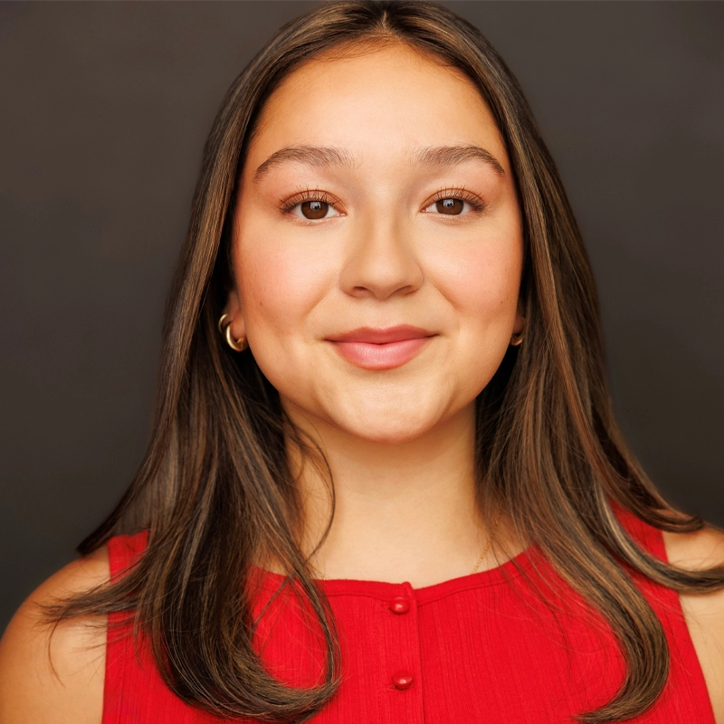Isa Hernandez, a young woman with straight brown hair and light skin, smiles softly at the camera. She wears a sleeveless red top with buttons and small gold hoop earrings, standing out against the plain dark background.