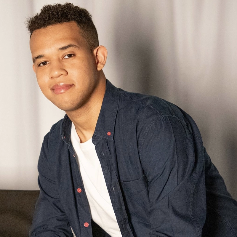 Juan Herrera, a young man with short curly hair, smiles softly while sitting slightly angled. He wears a dark blue button-up shirt over a white t-shirt. The softly lit, plain background creates a relaxed and casual atmosphere.