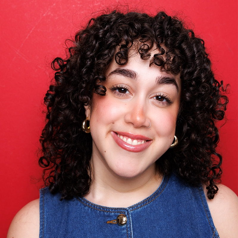 Ireyssa Cardona, with curly dark hair, gold hoop earrings, and a nose ring, smiles warmly at the camera. She wears a blue denim top with a button against a bright red background. Her makeup is natural, complementing her warm and friendly expression.
