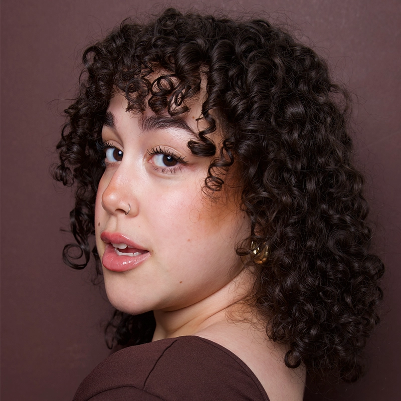 A person with short, dark, tightly curled hair looks over their shoulder. They have clear skin, a small nose ring, and gold hoop earrings—an effortlessly confident look reminiscent of Ireyssa Cardona. The background is a solid maroon color, complementing their maroon top.