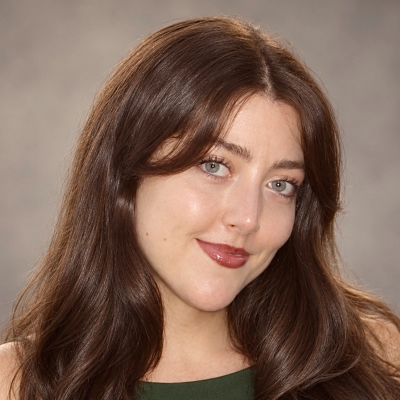 A woman with long, wavy brown hair smiles gently. She has light skin, blue eyes, and wears natural makeup with pink lipstick. Ireyssa Cardona is dressed in a sleeveless green top, and the background is a plain, softly blurred gray.
