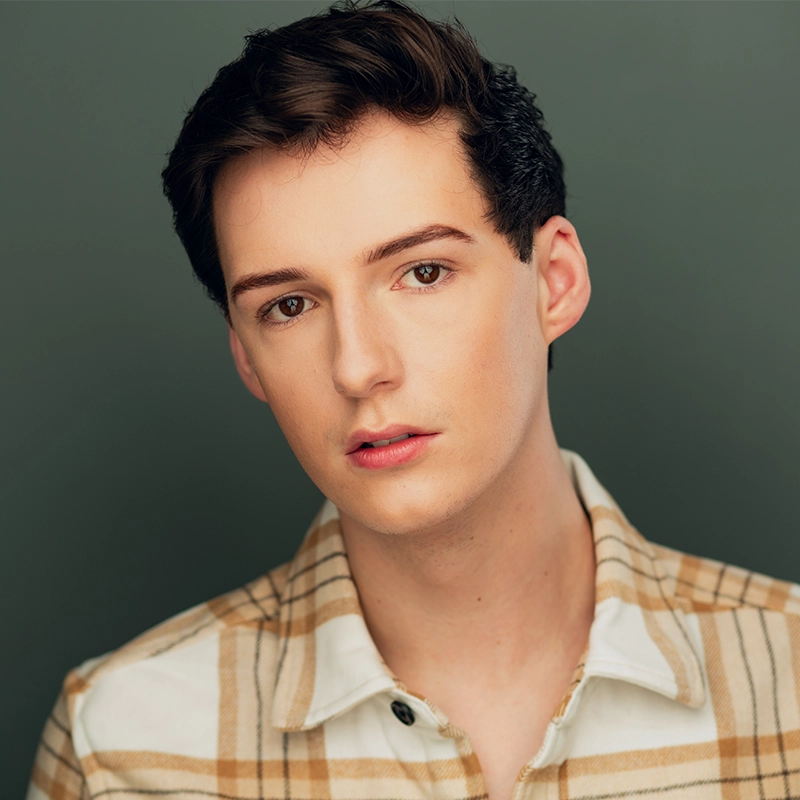 A young person with short, dark brown hair and fair skin looks at the camera with a neutral expression. They wear a beige and white plaid button-up shirt. The background is a muted green color. The lighting is soft and even.