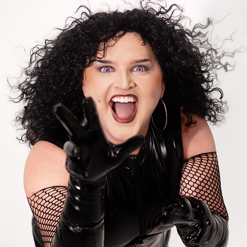 Aidan Michael Kelly, with curly black hair, dramatic eye makeup, and purple lipstick, poses energetically—reaching toward the camera in hoop earrings, black fishnet sleeves, shiny gloves, and a matching top against a white background.