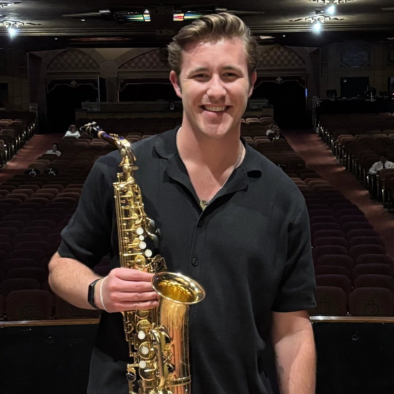 A smiling man, Alex Jashinski, in a black shirt holds a golden saxophone in both hands. He stands on stage in a theater with rows of red seats and ornate decor visible in the background, with just a few people seated far behind him.