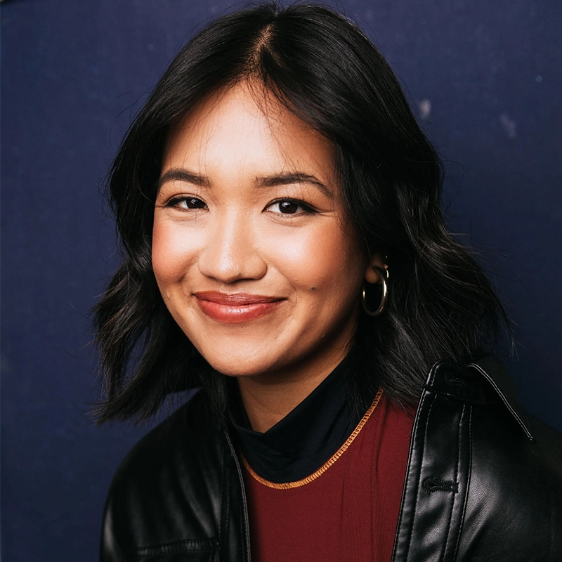 A young woman with wavy, shoulder-length black hair smiles warmly against a dark blue background. She wears gold hoop earrings, a burgundy top, a thin gold necklace, and a black leather jacket. Her makeup is natural, highlighting her dimples.