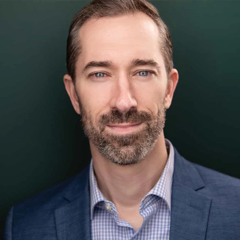 Kenna Wilson stands facing forward, smiling slightly. He has short brown hair, a neatly trimmed beard, and wears a blue blazer over a light checkered shirt. The professional, studio-lit portrait features a dark green background.